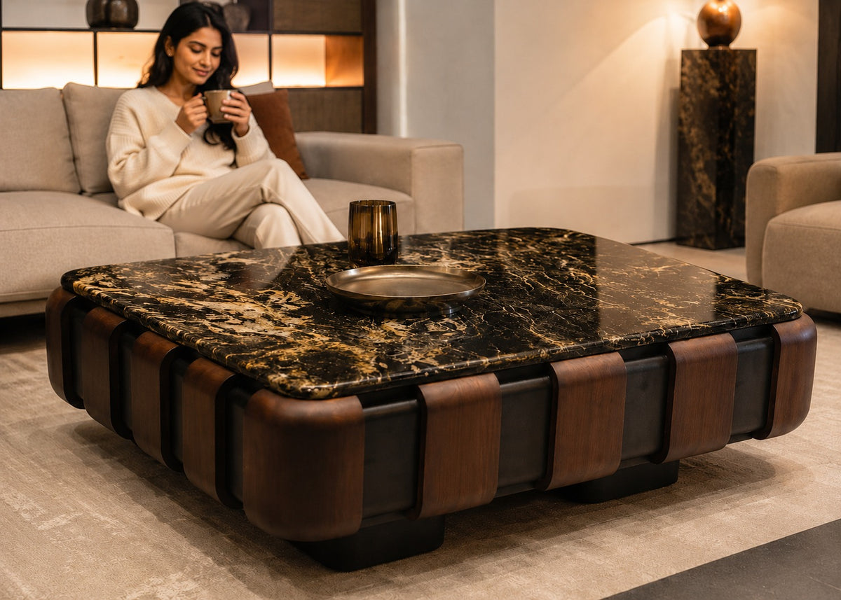 A modern square coffee table with a black and gold marble top and brown leather-like side panels, shown in a styled living room. A woman sits on a beige sofa behind the table, holding a cup.