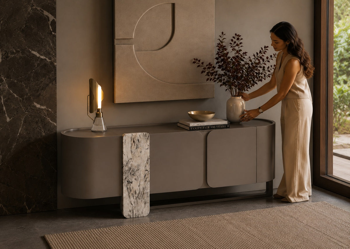 A woman in a beige sleeveless dress stands beside a modern curved gray console table in a styled interior. The table has a vertical white-and-gray marble side panel, a lit wall lamp, books, a bowl, and a vase with dried branches.