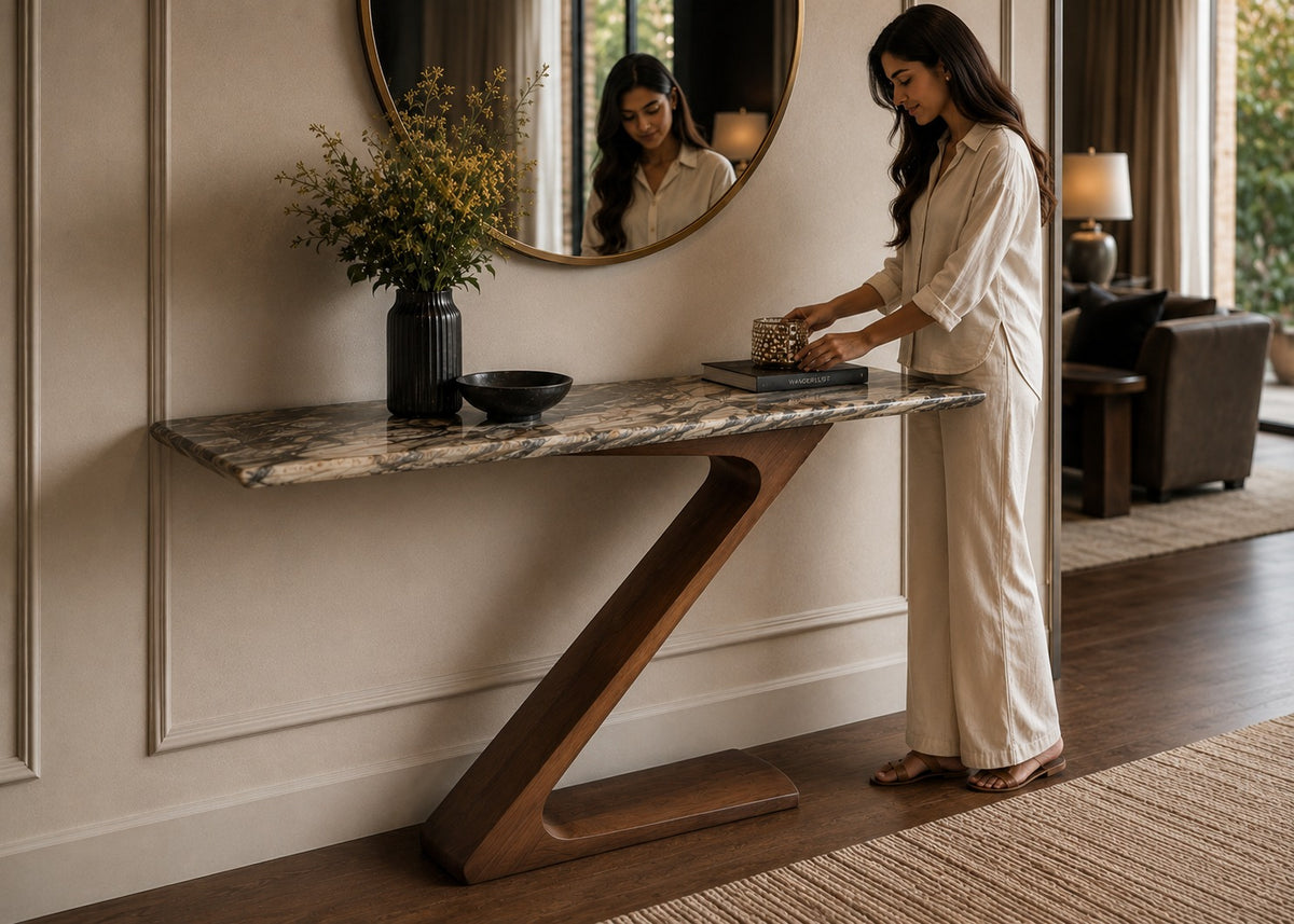 A woman in a cream-colored robe stands beside a modern console table with a marble top and sculptural wooden base in a styled interior room.