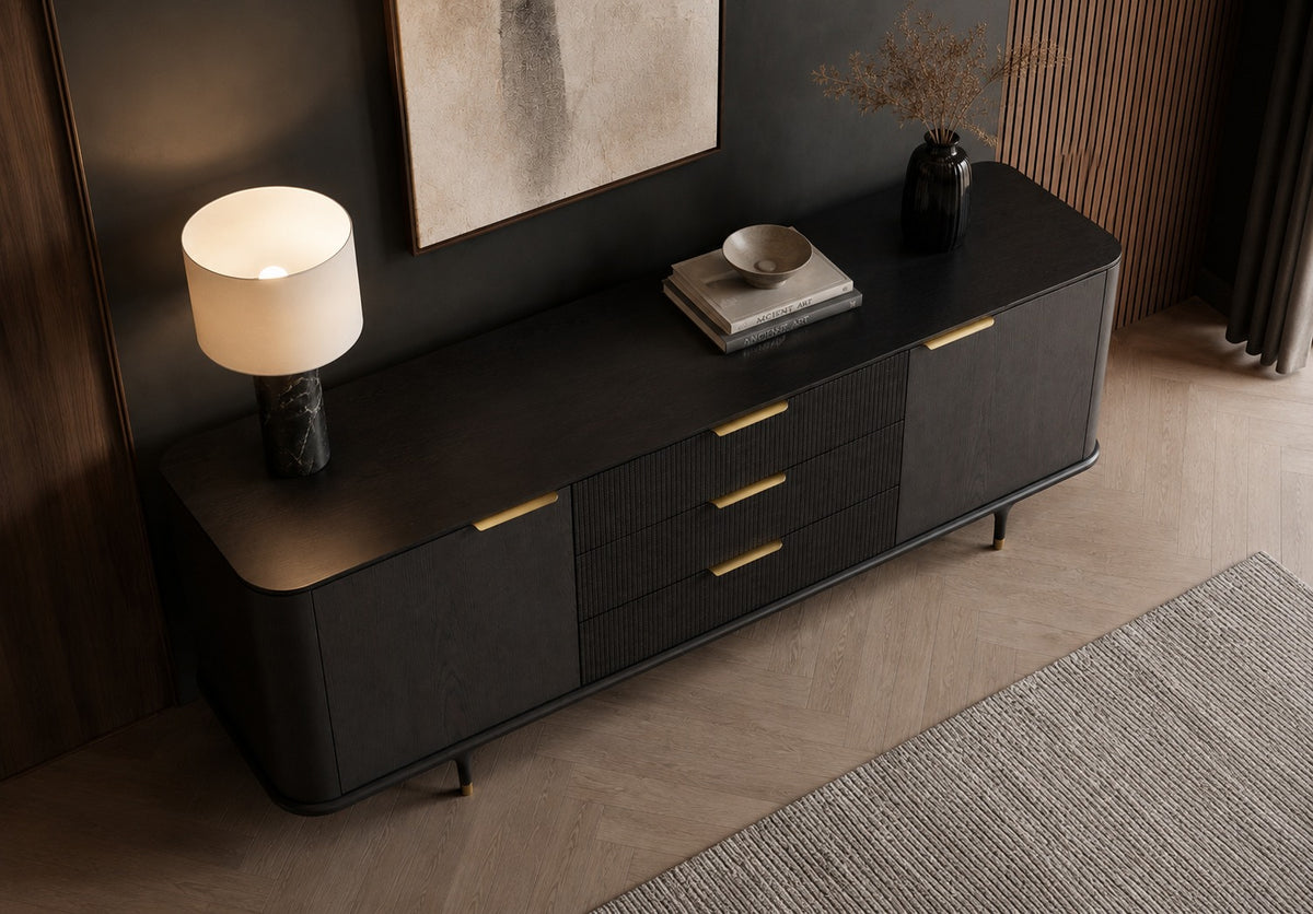 A modern black sideboard cabinet with gold-tone handles and legs, styled in a contemporary room with a lit table lamp, books, and a bowl on top.