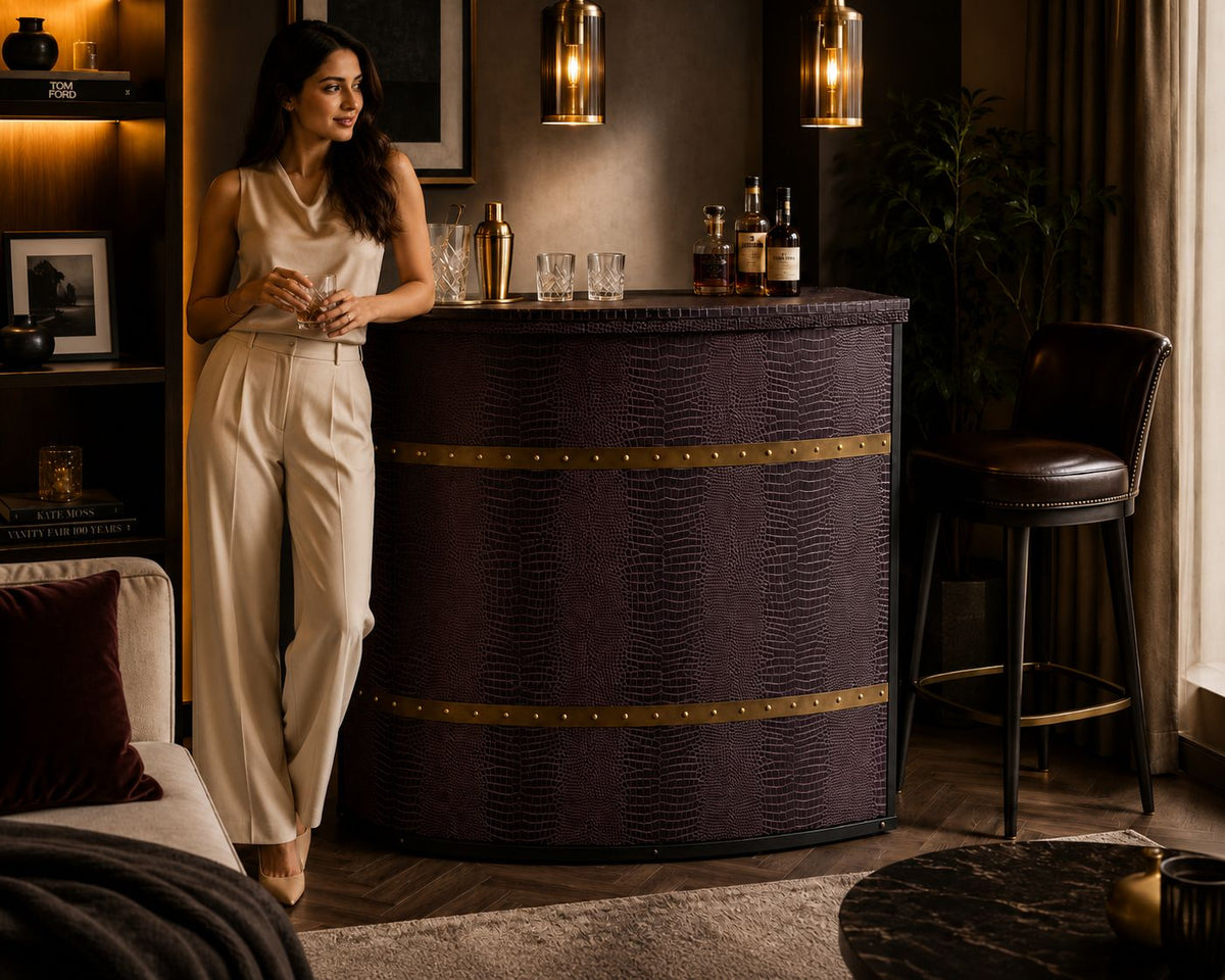 A woman in a cream satin sleeveless top and wide-leg trousers stands beside a curved bar counter with a dark textured finish and gold-toned trim in a warmly lit lounge setting.