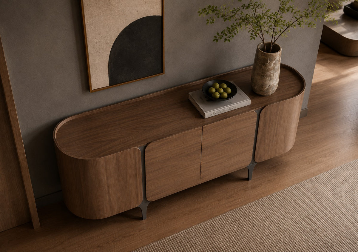 A modern curved wooden sideboard or console cabinet in a warm medium brown wood finish, styled in a minimalist room with a textured gray wall, abstract wall art, a ceramic vase with greenery, and a bowl of green olives on top.