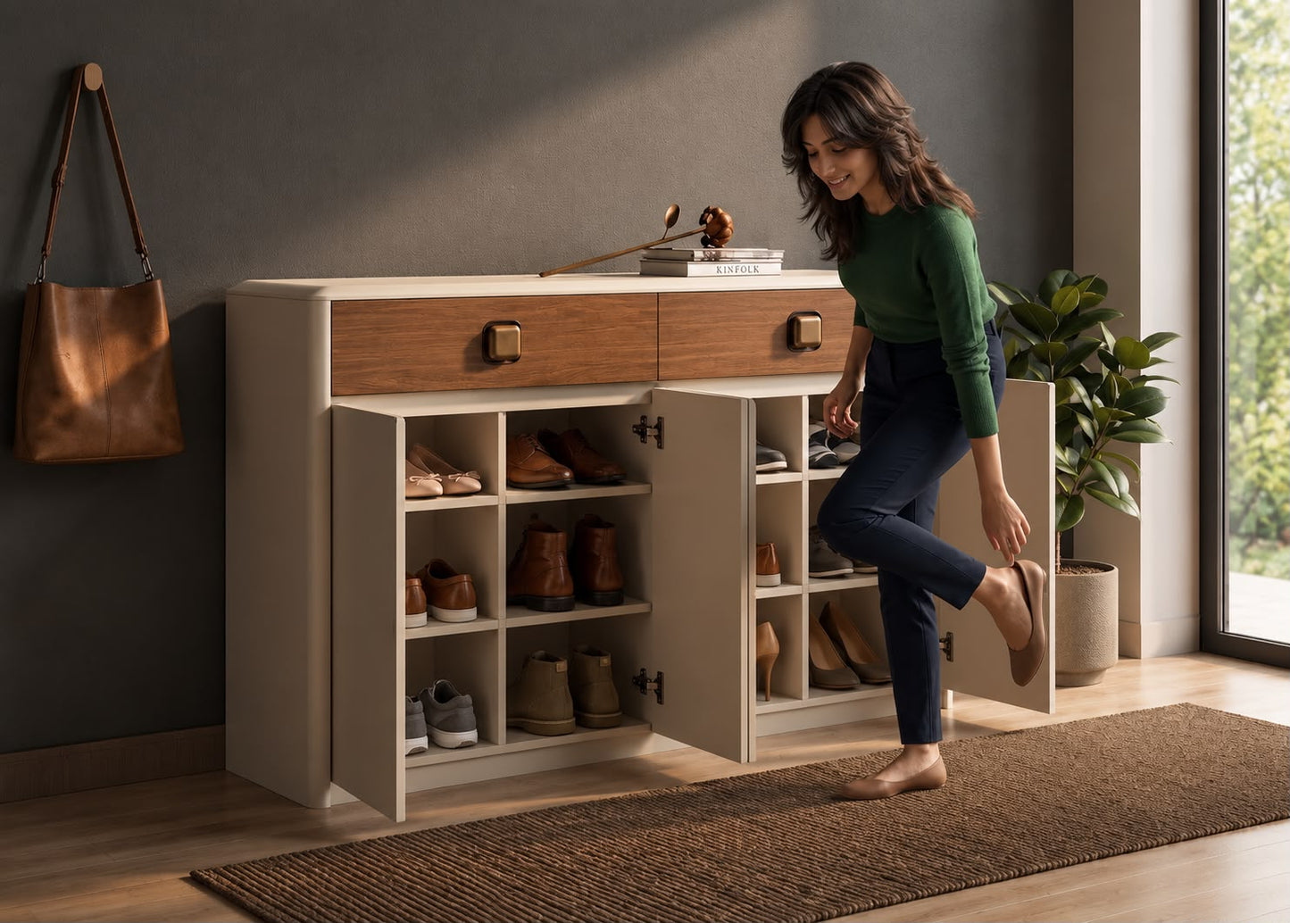 A woman in a green top and dark pants is barefoot, putting on a shoe while standing next to an open cream and wood shoe cabinet in a modern room.