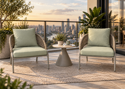 Two outdoor wicker armchairs with pale sage green cushions are arranged on a woven rug with a small round side table between them on a balcony overlooking a city skyline at sunset.