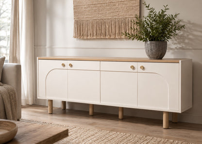 A modern sideboard cabinet in white with a light wood top and legs, styled in a bright living room next to a sofa with a potted plant on top.