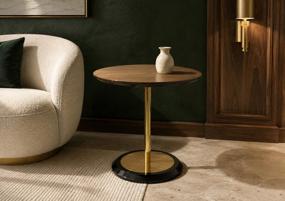 Round wooden side table with gold base next to a beige armchair in a room with dark walls.