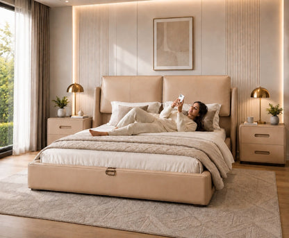 A woman relaxes on a beige upholstered bed in a modern, warm-toned bedroom. The bed has a matching headboard and footboard, with cream bedding, bedside tables, gold table lamps, and a light wood floor.