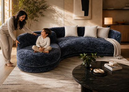 A curved plush sectional sofa in deep navy blue textured fabric, styled in a warm, modern living room with a woman and a child interacting with it.