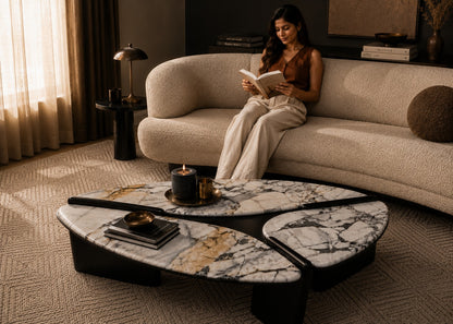 Woman reading a book on a beige sofa in a dimly lit room with a marble coffee table.