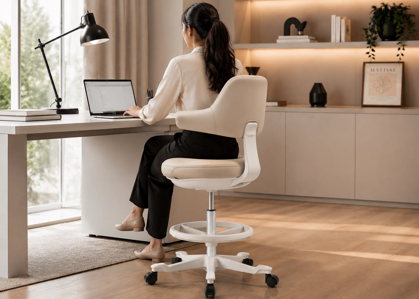A woman sits on a white swivel office chair at a light wood desk in a bright, modern home office, using a laptop. The chair has a curved backrest, padded seat, and a white five-star wheeled base.