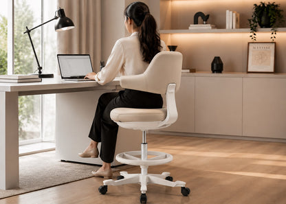 A woman sits on a white swivel office chair at a light wood desk in a bright, modern home office, using a laptop. The chair has a curved backrest, padded seat, and a white five-star wheeled base.