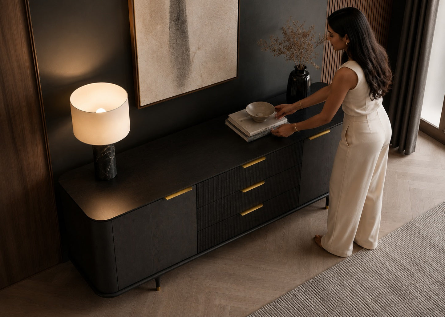 A woman in a cream sleeveless jumpsuit stands beside a dark wood sideboard in a modern room, placing a bowl on a stack of books. The sideboard has gold-tone handles and a lit table lamp on top.