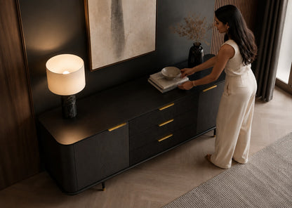 A woman in a cream sleeveless jumpsuit stands beside a dark wood sideboard in a modern room, placing a bowl on a stack of books. The sideboard has gold-tone handles and a lit table lamp on top.