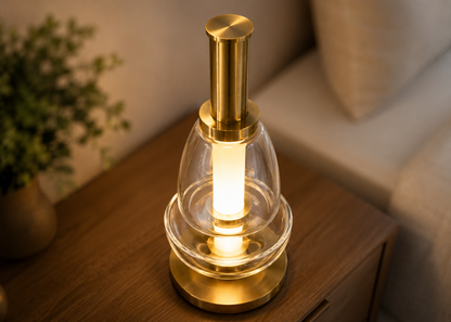 A modern table lamp with a brushed brass cylindrical top and base, a clear glass dome shade, and a glowing warm white vertical bulb, shown on a wooden bedside table in a softly lit room.