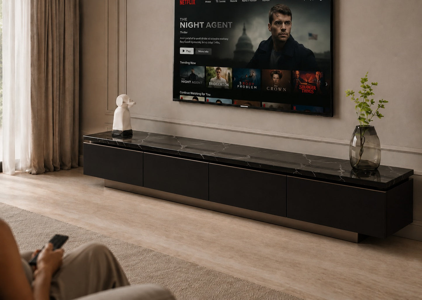 A modern black floating TV console with a dark marble-look top, shown in a living room setting below a wall-mounted television. A person is seated in the foreground holding a remote, and decorative objects including a small white sculpture and a glass vase with greenery sit on the console.