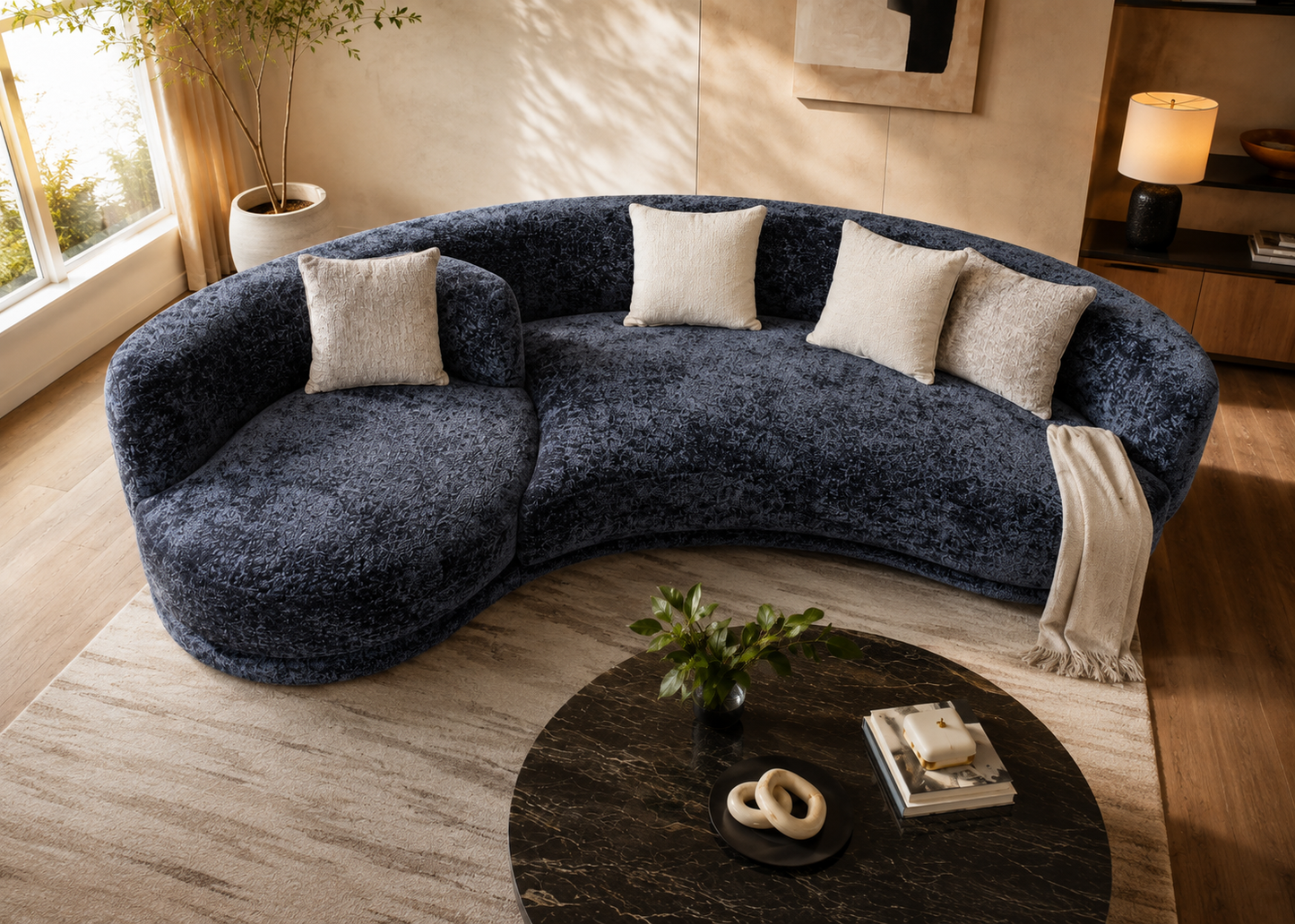 A curved, plush sectional sofa upholstered in dark heathered blue-gray fabric, styled in a warm, modern living room with cream throw pillows, a beige rug, and a round dark coffee table.