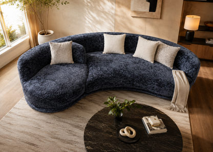 A curved, plush sectional sofa upholstered in dark heathered blue-gray fabric, styled in a warm, modern living room with cream throw pillows, a beige rug, and a round dark coffee table.