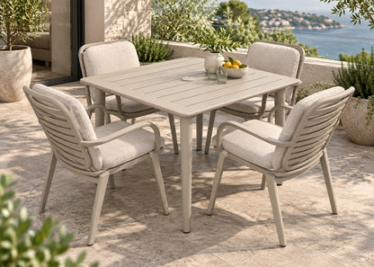 A 5-piece outdoor patio dining set featuring a square slatted table and four armchairs with light beige cushions, set on a stone patio overlooking a scenic coastal view.
