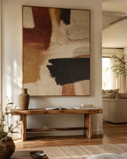 A large abstract wall art piece with sweeping brushstrokes in beige, brown, tan, black, and cream hangs above a rustic wooden bench in a softly lit living room. A terracotta vase and books sit on the bench, with a sofa and plants visible in the background.