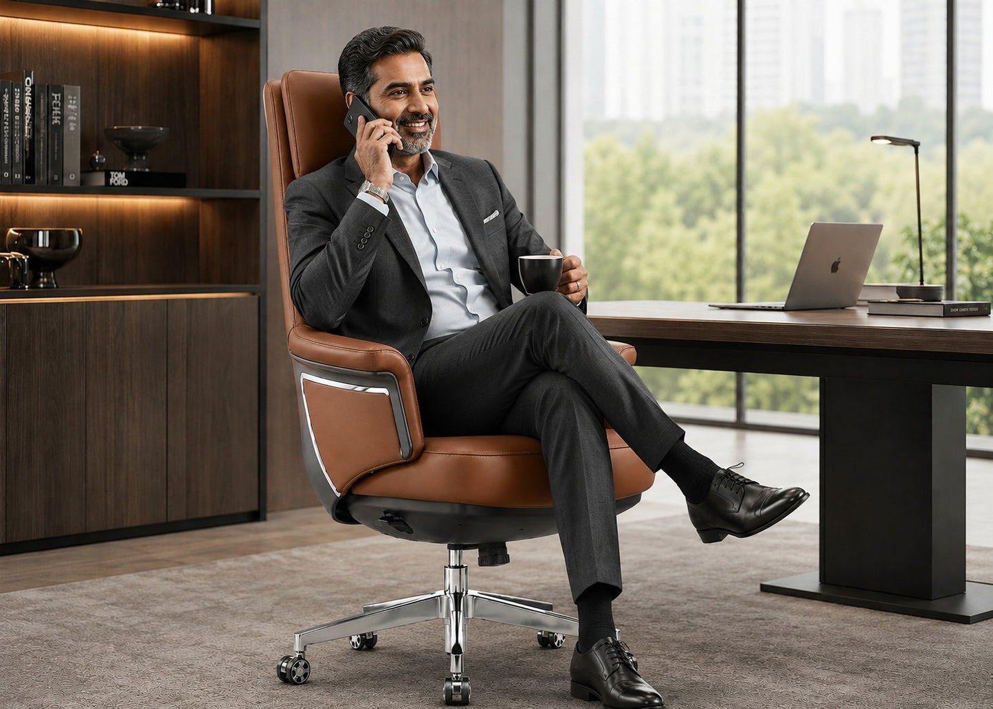 A man in a dark suit sits relaxed in a brown leather office chair with a chrome base, holding a phone and a mug in a modern office setting with a desk and large windows.