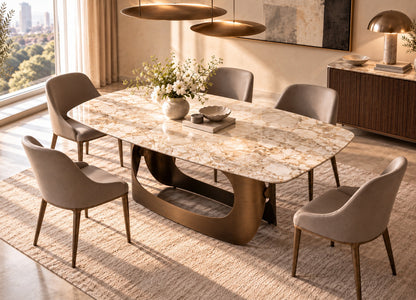 An oval dining table with a polished beige marble top and a sculptural bronze base, surrounded by six upholstered dining chairs in a warm, modern dining room setting.