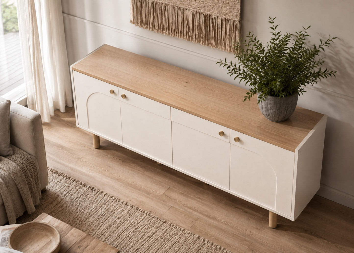A modern white sideboard cabinet with a light natural wood top, styled in a bright living room next to a sofa with a potted green plant on top.