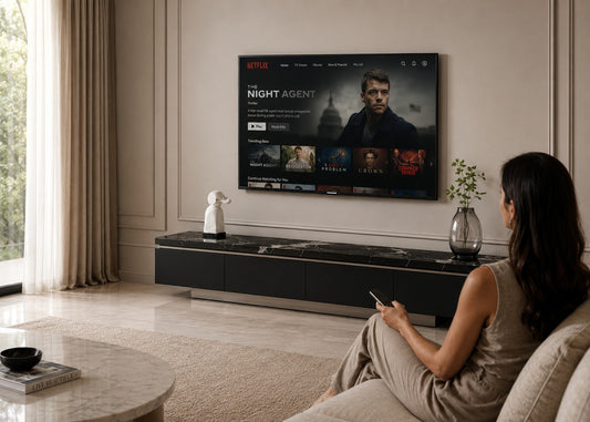 A woman sits on a beige sofa in a modern living room, watching a large wall-mounted TV displaying the Netflix streaming interface. The room has cream walls, a black marble media console, and a round marble coffee table.