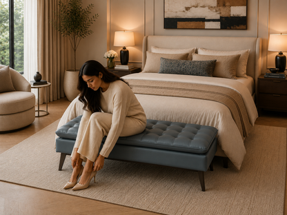 Woman sitting on a blue ottoman in a bedroom with beige bedding and decor.