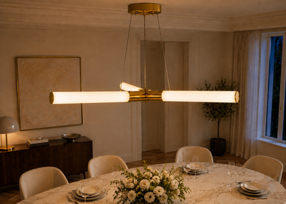 A modern linear chandelier with two horizontal frosted glass tubes and a brushed gold central bar, hanging over a marble dining table in a warmly lit, elegant dining room.