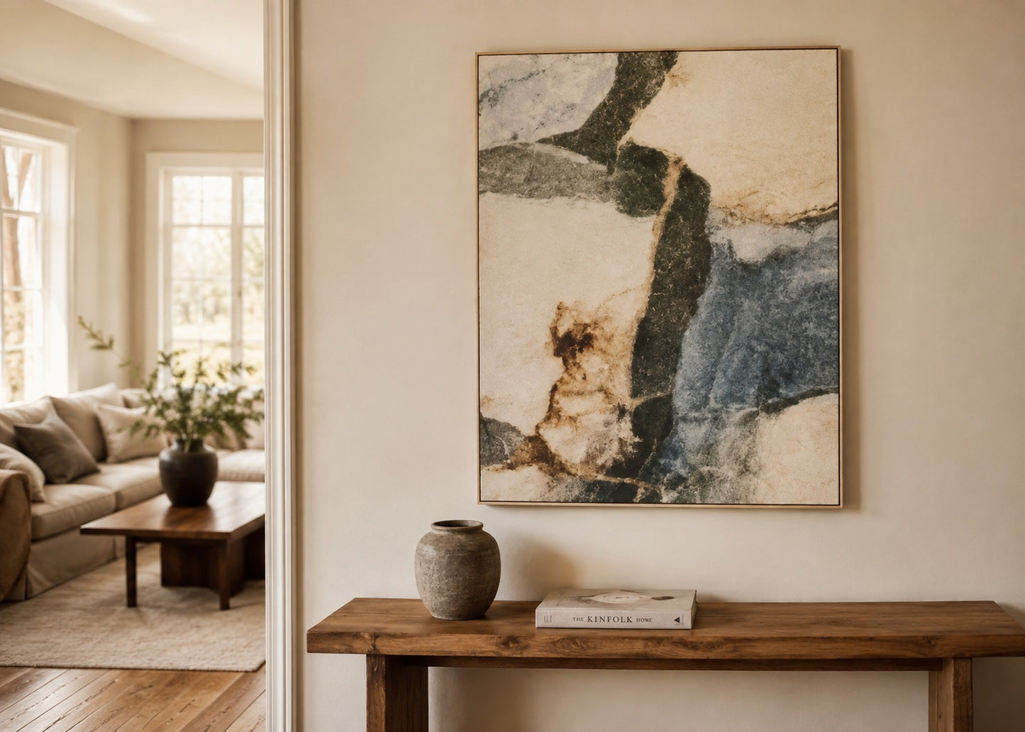 An abstract framed wall art print hangs on a beige wall above a rustic wooden console table in a styled living room interior. The artwork features flowing brushstrokes of cream, slate blue, charcoal, and tan.