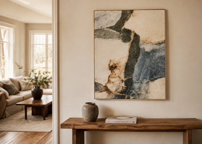 An abstract framed wall art print hangs on a beige wall above a rustic wooden console table in a styled living room interior. The artwork features flowing brushstrokes of cream, slate blue, charcoal, and tan.