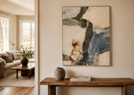 An abstract framed wall art print hangs on a beige wall above a rustic wooden console table in a styled living room interior. The artwork features flowing brushstrokes of cream, slate blue, charcoal, and tan.