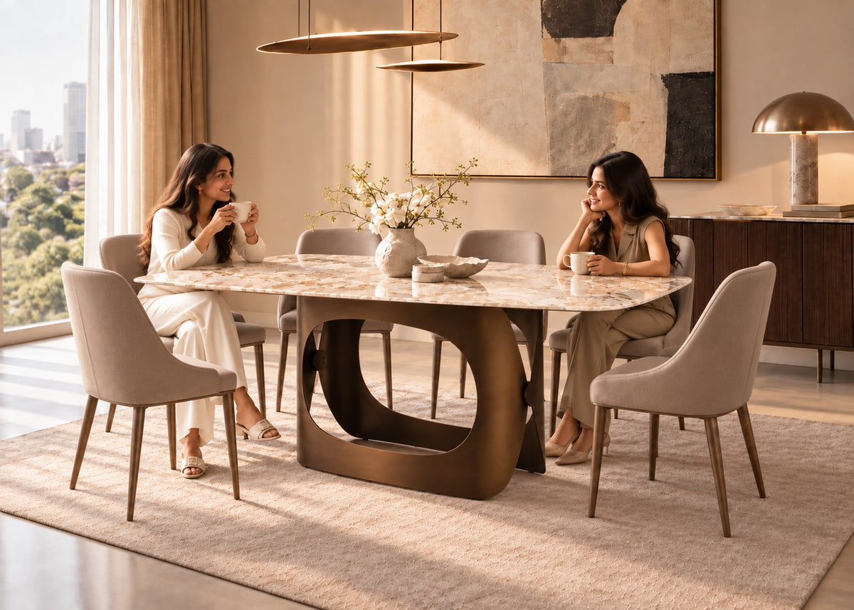 A modern dining room scene with two women seated at a rectangular marble-top dining table with a sculptural bronze base, surrounded by upholstered dining chairs. The room has warm neutral tones, large windows, pendant lights, and wall art.