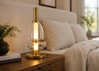 A modern table lamp with a polished brass base and neck, a clear glass body, and a glowing warm white vertical light, placed on a wooden bedside table next to a bed in a softly lit bedroom.