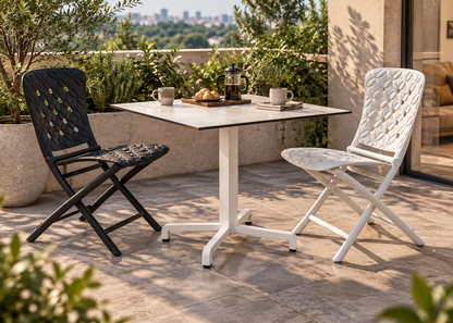 Two modern outdoor folding chairs and a matching square table on a patio, with one chair in black and one in white, set against a backdrop of greenery and a distant city skyline.