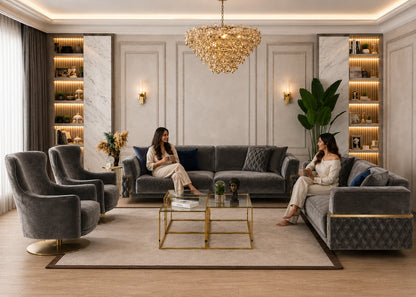 A luxury living room scene featuring a modern gray velvet sectional sofa, two matching armchairs, a glass-top coffee table with gold legs, and two women seated on the sofa in a highly styled interior with a crystal chandelier and built-in shelves.