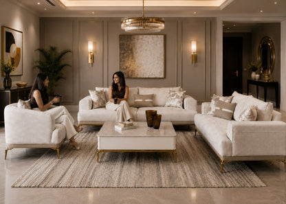 A luxury living room featuring a modern sectional sofa, matching armchair, and a rectangular coffee table, all upholstered in a light cream fabric with gold legs. Two women are seated in the furniture, and the room is decorated with a large textured area rug, wall sconces, and a gold chandelier.