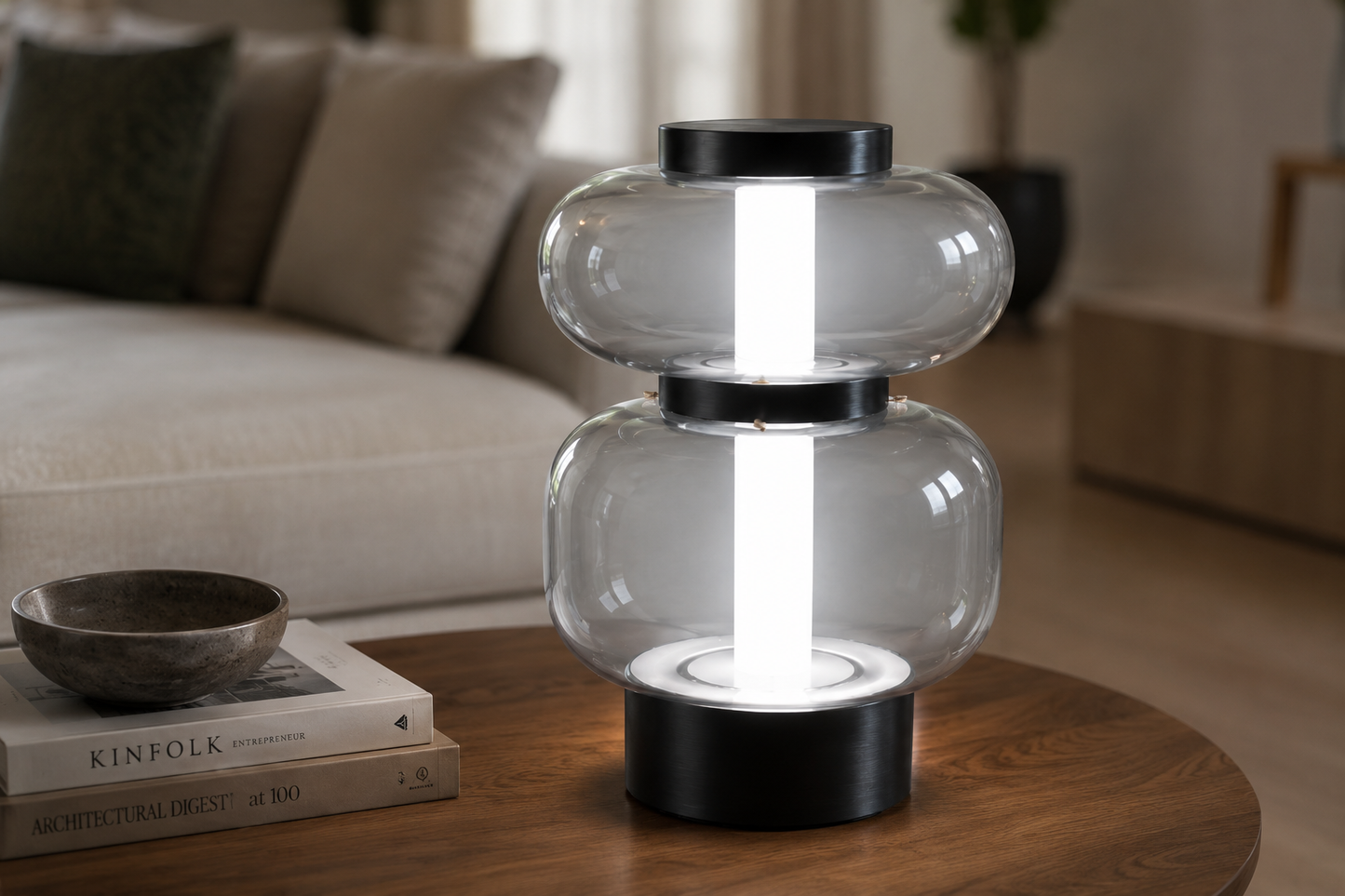 A modern two-tier table lamp with clear glass shades and a matte black base, shown lit on a round wooden table in a living room setting.