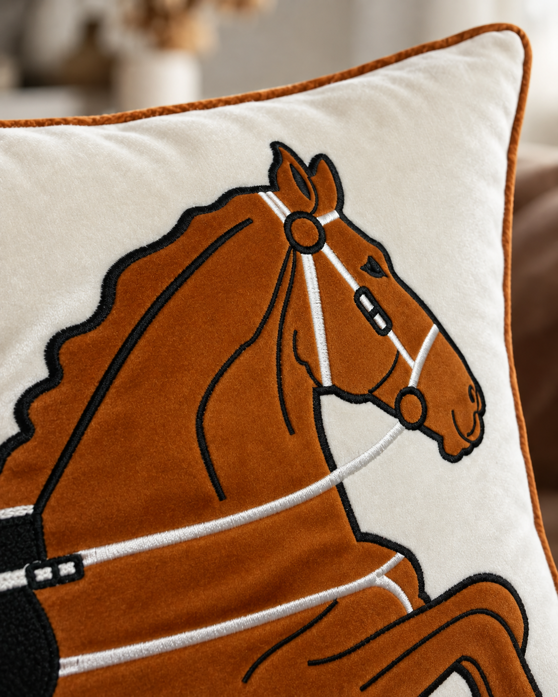 Close-up of a decorative throw pillow featuring an embroidered brown horse design with white reins and black outlines on a cream background, with a rust-brown piped edge.