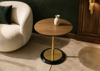 Round wooden side table with gold base next to a beige armchair in a room.