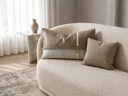 A curved upholstered sofa in a warm beige bouclé fabric, styled with three neutral-toned throw pillows in a softly lit living room. A small marble side table with a vase stands near the window in the background.