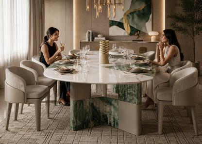 A luxury round dining table with a white marble top and green marble base, surrounded by beige upholstered chairs, with two women seated at the head of the table in a modern, elegantly decorated dining room.