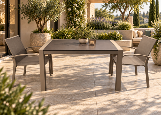 A rectangular outdoor dining table with a dark gray top and gray frame, accompanied by two matching armchairs on a sunlit patio. The setting features a modern house exterior, large potted olive trees, and a stone seating area in the background.