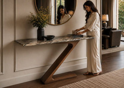 A woman in a cream-colored robe stands beside a modern console table with a marble top and sculptural wooden base in a styled interior room.