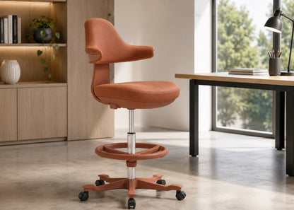 An orange-brown upholstered swivel office chair with a circular footrest and black caster wheels, shown in a modern home office setting next to a desk by a window.