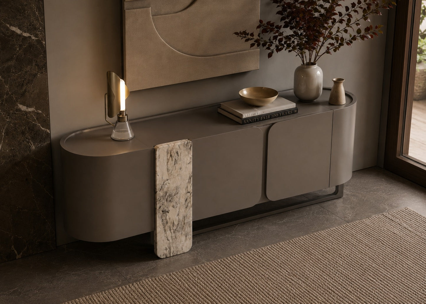 A modern curved sideboard or credenza in a warm taupe-gray finish, styled in a contemporary interior with a marble-look vertical panel leaning against it, a lit sculptural table lamp, books, a bowl, and vases near a window.