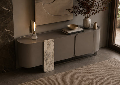 A modern curved sideboard or credenza in a warm taupe-gray finish, styled in a contemporary interior with a marble-look vertical panel leaning against it, a lit sculptural table lamp, books, a bowl, and vases near a window.
