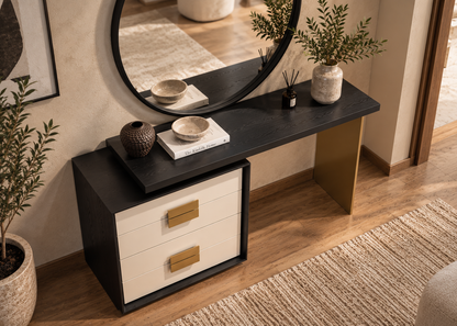 A modern two-drawer console table with a black top and frame, cream drawer fronts, and gold-tone handles, styled in a warm-toned room with a round mirror, plants, and decorative objects.