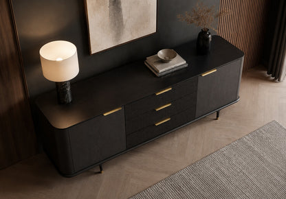 A modern black sideboard cabinet with gold-tone handles and legs, styled in a contemporary room with a lit table lamp, books, and a bowl on top.
