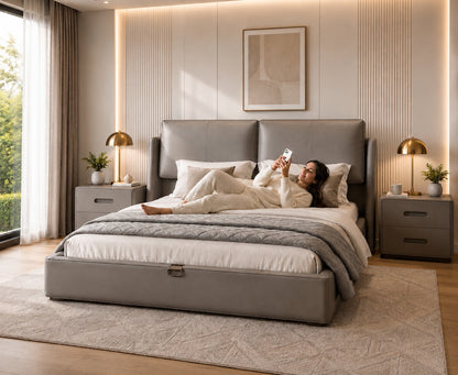 A woman relaxes on a modern gray upholstered bed in a styled bedroom. The bed has a low storage footboard, white bedding, and a gray throw blanket.